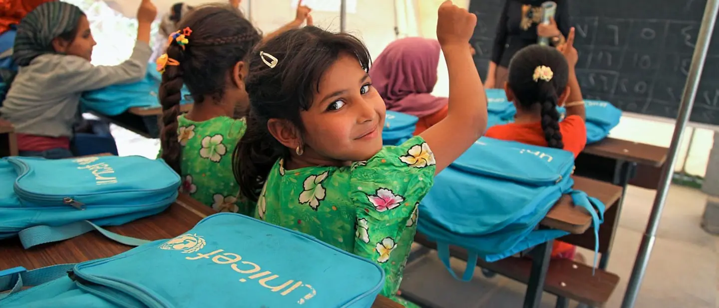 UNICEF setzt sich in rund 190 Ländern mit Hilfsprojekten für Kinder ein | © UNICEF/UNI194536/Khuzaie Kinder sitzen in einem Klassenzimmer und melden sich. Vor ihnen liegen Schulrucksäcke von UNICEF.