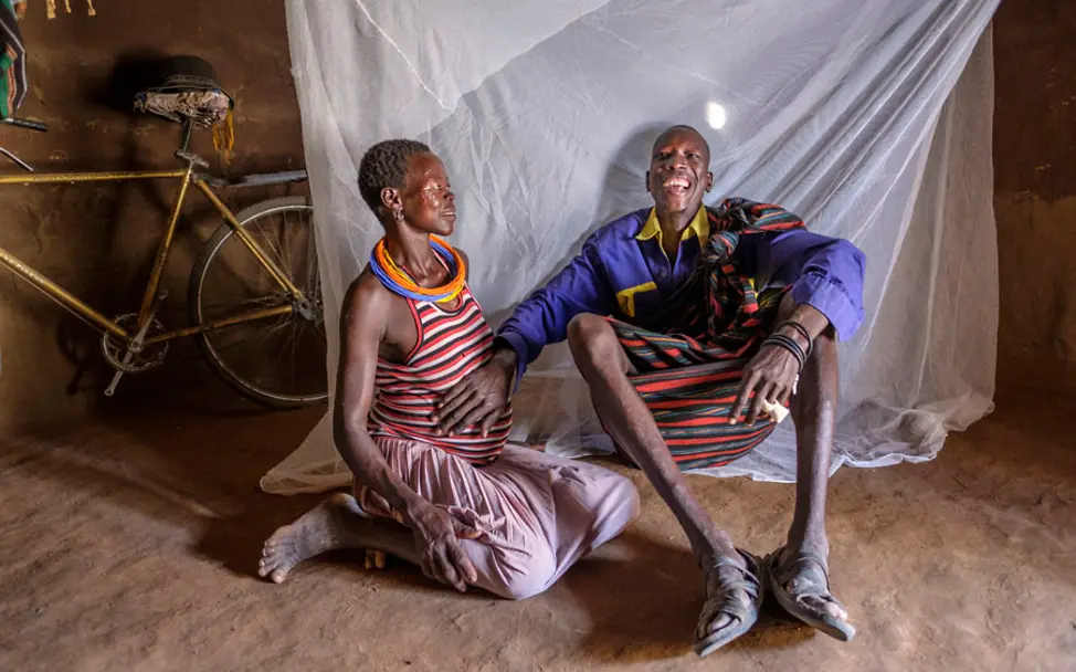 Loli mit einer seiner vier Frauen - sie erwartet sein zwanzigstes Kind | © UNICEF/Ose Uganda: Loli sitzt lachend neben einer seiner vier Frauen