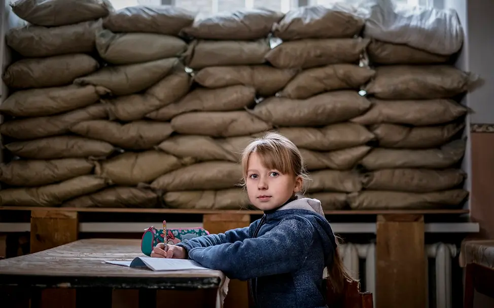 Ein Mädchen in der Sch| © UNICEF/UN0150817/Gilbertson VII Photo Ein Mädchen sitzt in einem Klassenzimmer an einem Pult. Im Hintergrund verdecken Sandsäcke das Fenster.