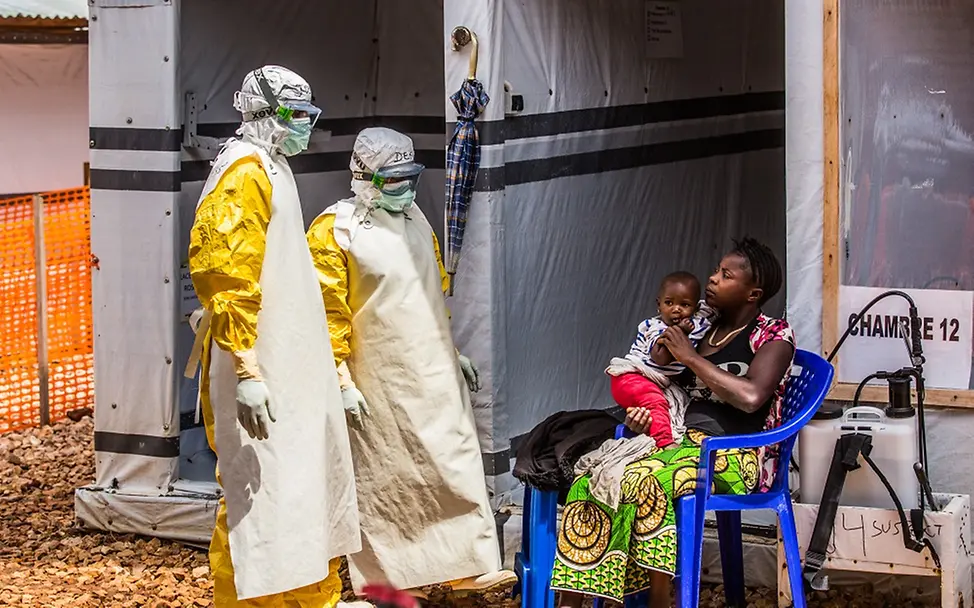 Zwei Gesundheitshelfer besuchen Mutter und Tochter in der Quarantäne-Zone des Ebola Behandlungszentrums in Butembo. | © UNICEF/Tremeau Zwei in Schutzkleidung gehüllte Gesundheitshelfer reden mit einer sitzenden Frau, die ihre Tochter auf dem Schoß hält.
