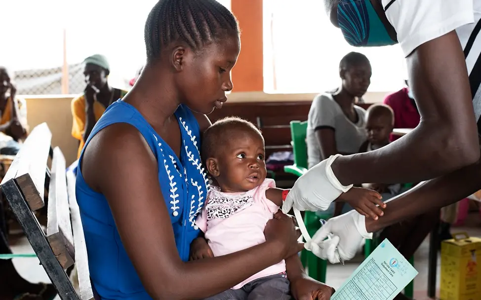 Eine Gesundheitshelferin untersucht Flora auf akute Mangelernährung. | © UNICEF/Ryeng Flora sitzt auf dem Schoss ihrer Mutter, während eine Gesundheitshelferin ihren Armumfang misst.