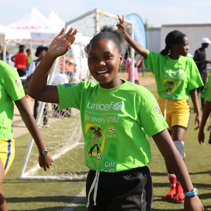 Mit Sport gegen Aids: Das UNICEF-Fußballprogramm in Namibia richtet sich an benachteiligte Mädchen. | © UNICEF/Mutseyekwa Ein Mädchen aus einem Fußball-Team winkt fröhlich in die Kamera. Sie trägt ein UNICEF-Trikot.