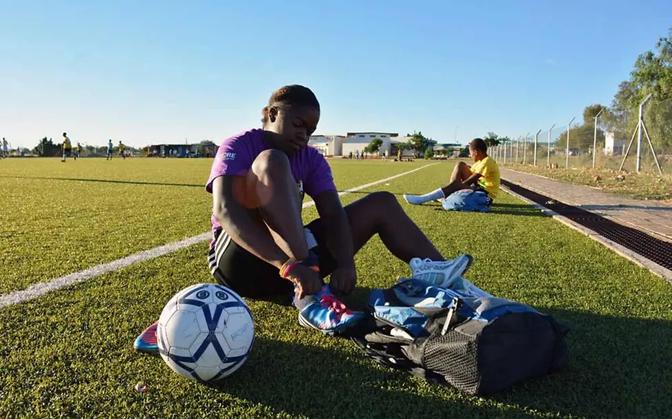 UNICEF stattet die Mädchen in Namibia mit Sportequipment aus. | © UNICEF/NFA Ein Mädchen sitzt auf einem Fußballfeld und schnürt ihre Fußballschuhe.