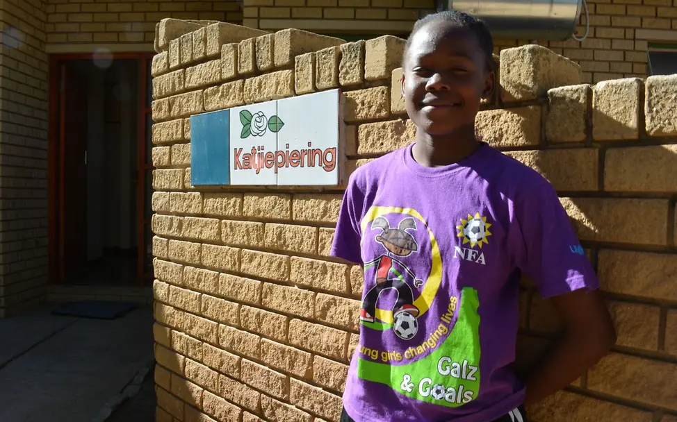 Das UNICEF-Programm 'Mädels & Ziele' verändert das Leben der Mädchen. | © UNICEF/NFA Ein Mädchen in einem lila Trikot steht lächelnd an einer Wand aus Ziegelsteinen.