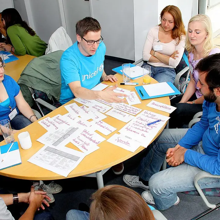 Menschen sitzen um einen Tisch, auf dem Formulare liegen und sprechen mit Mitgliedern der UNICEF Hochschulgruppe.