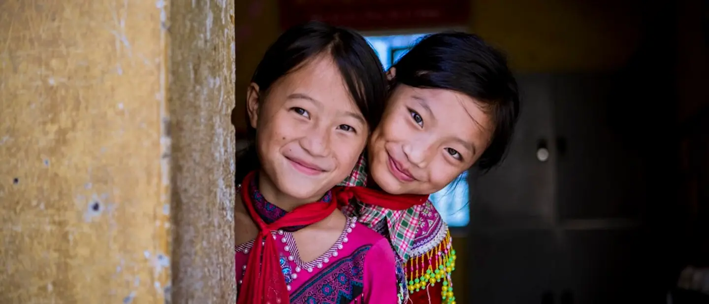 Viertnam: Zwei Mädchen gucken hinter einer Mauer hervor und lächeln.
