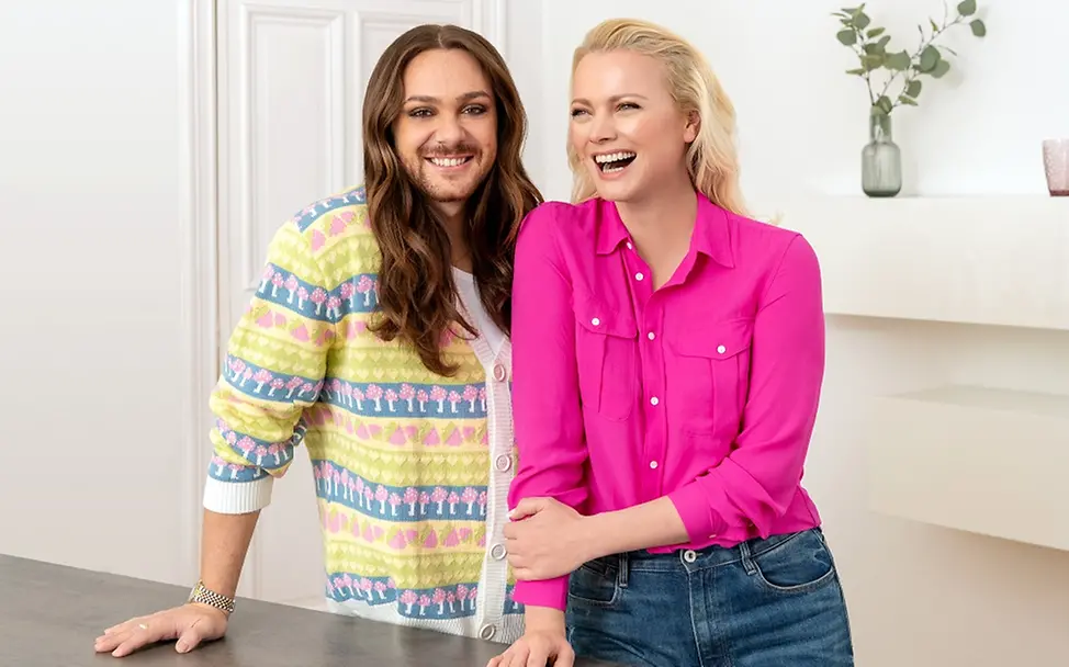 Franziska Knuppe und Riccardo Simonetti stehen vor einem Tisch nebeneinander und lächeln.