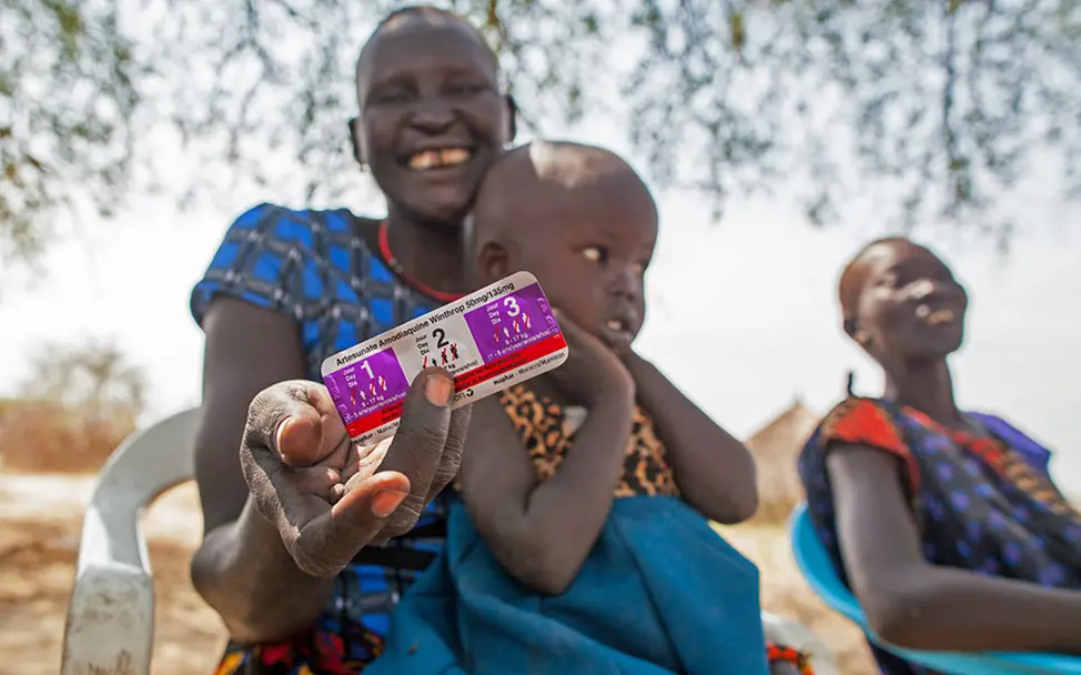Eine lächelnde Mutter im Südsudan zeigt das Malaria-Medikament für ihre infizierte Tochter.