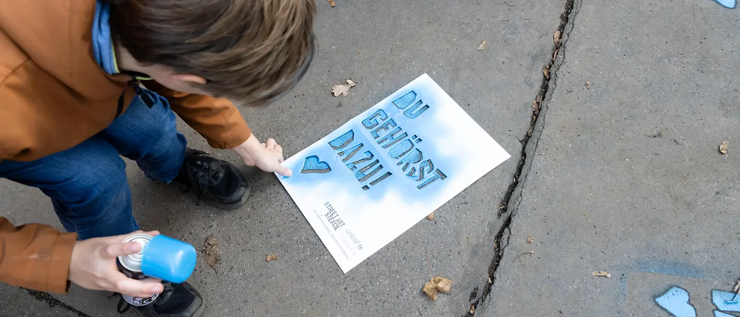 Ein Junge sprüht mit einer Sprühdose "Du gehörst dazu!" auf den Steinboden. 