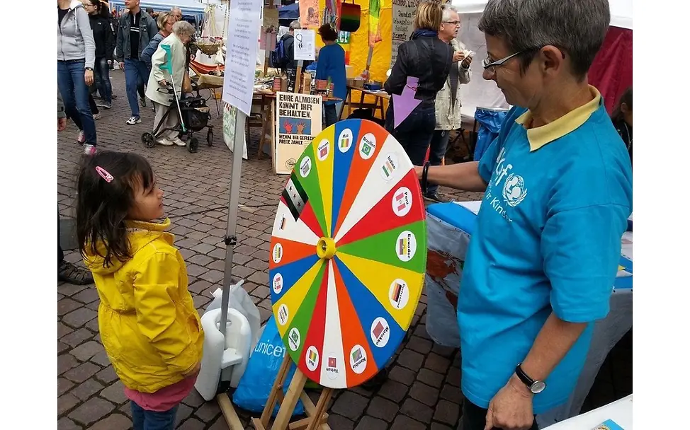 Unser Schicksalsrad beim Markt der Kulturen Ein Mädchen und eine Ehrenamtliche von UNCICEF schauen auf das bunte Schicksalsrad (Glücksrad) mit Länderflaggen