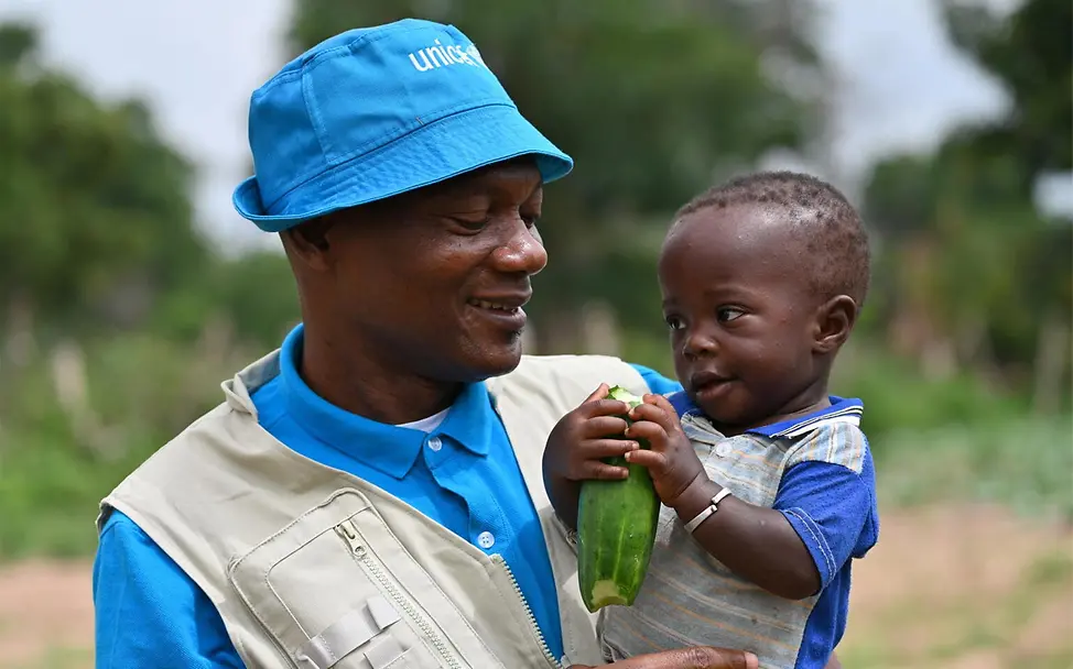Hungersnot verhindern: Ein Kleinkind mit Zucchini in der Hand bei einem UNICEF-Helfer auf dem Arm