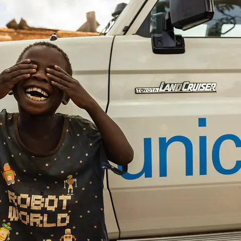 Richtig Spenden: Ein Junge steht vor einem Auto mit UNICEF-Logo und hält sich lachend die Augen zu.