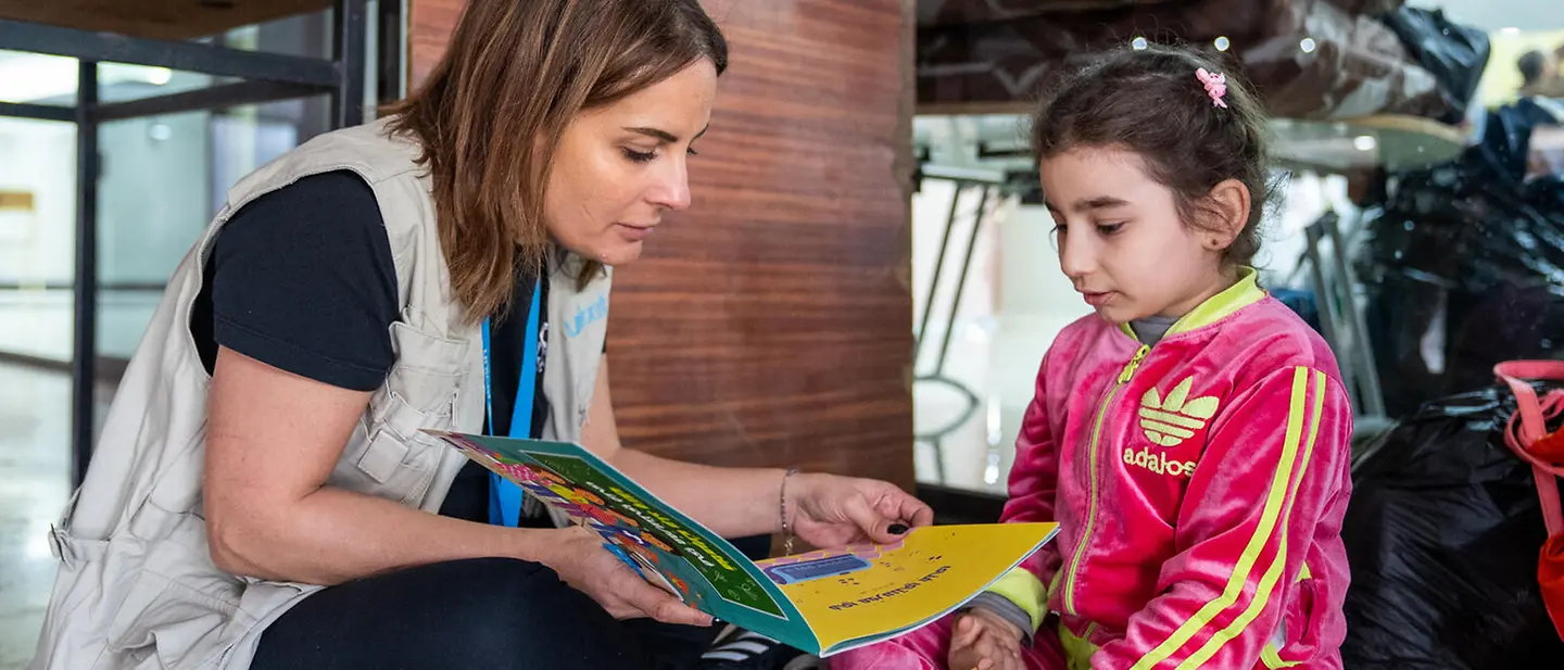 Libanon spenden: Eine UNICEF-Helferin schaut mit einem Mädchen in einer Notunterkunft ein Buch an 
