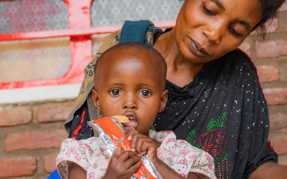 Kind sitzt auf dem Schoß der Mutter und erhält therapeutische Nahrung in Shasha