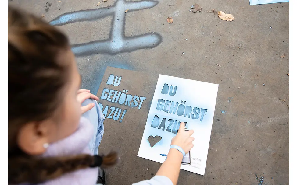 Du gehörst dazu: Botschaft wird am Boden markiert | © UNICEF/Etges Ein Mädchen sprüht mit einer Schablone den Schriftzug "du gehörst dazu" am Boden mit Sprayfarbe auf.