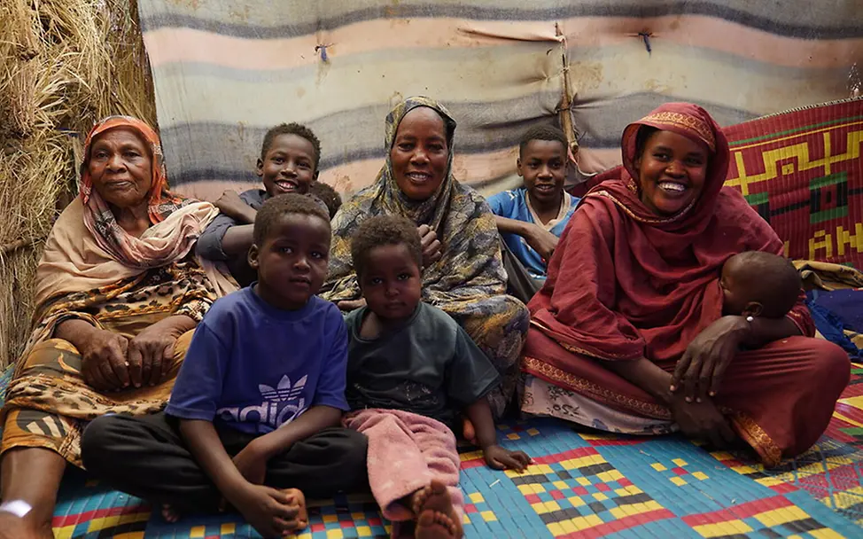 Sudan: Mehrere Kinder und drei Frauen sitzen gemeinsam in einer provisorischen Unterkunft auf Matten.