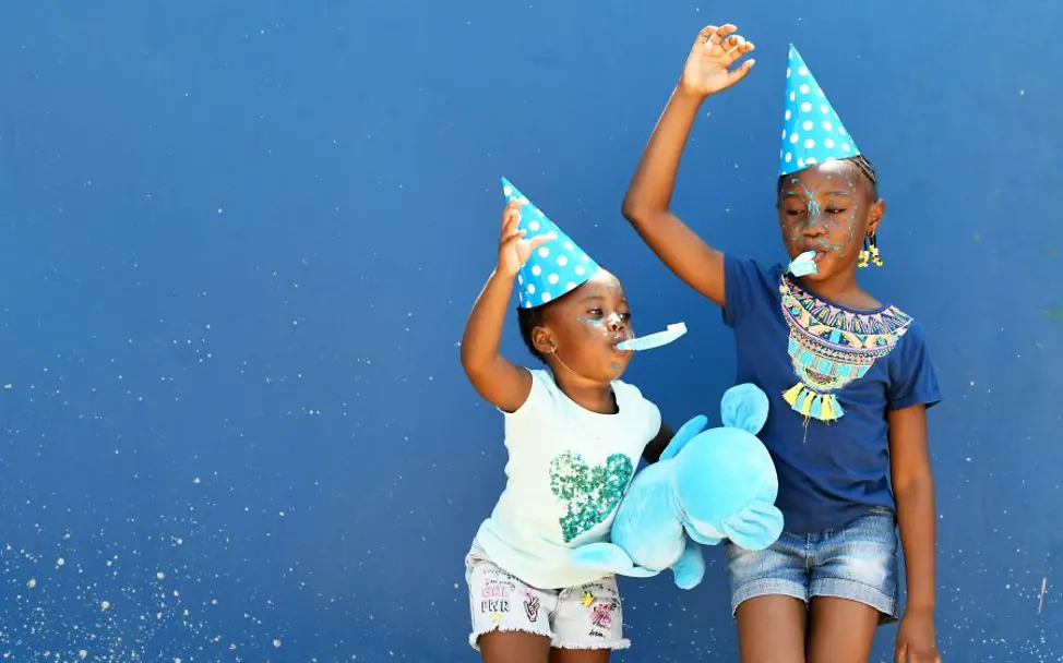 Kinderfreundliche Kommunen: Zwei Kinder stehen vor einer blauen Wand, tragen Partyhüte und halten ein großes Plüschtier.