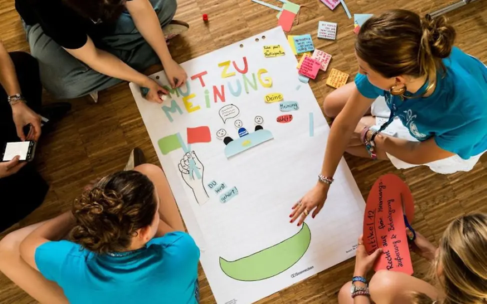 Yes-Day: Gruppe arbeitet sitzend an einem großen Plakat. | © UNICEF/UN0875915/Sachse-Grimm Schulaktion zum Tag der Kinderrechte: Jugendliche sitzen am Boden und arbeiten gemeinsam an einem großen Plakat.