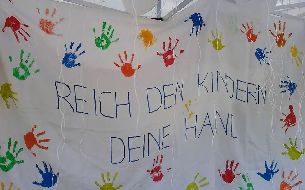 Auf einem Stoffbanner steht: "Reich den Kindern Deine Hand" mit vielen bunten Handabdrücken