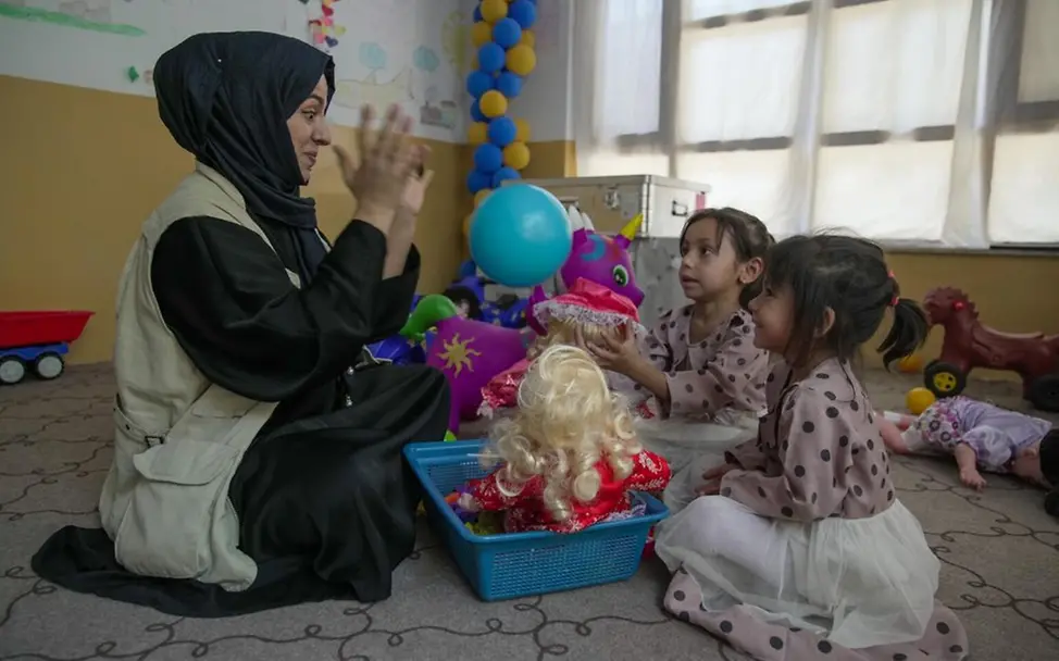 Eine UNICEF-Helferin spielt während einer psychosozialen Betreuung mit zwei Kindern. | © UNICEF/Karimi Eine UNICEF-Helferin sitzt mit zwei Kindern auf dem Boden. Sie spielen mit einem Ball und einer Puppe.