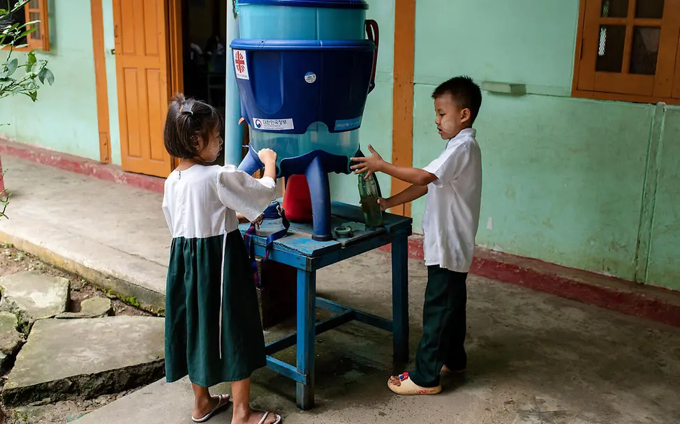 Sicheres Wasser hilft, die Ausbreitung von gefährlichen Krankheiten zu vermeiden. | © UNICEF/Ring Was ist Hunger einfach erklärt: Zwei Kinder stehen auf dem Schulhof an einem mobilen Wasserspender.