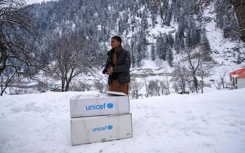 Im Osten Afghanistan erhält ein Jugendlicher warme Winterkleidung. | © UNICEF/Fazel Ein Junge steht im Schnee. Vor ihm stehen zwei UNICEF-Hilfspakete mit Winterkleidung.