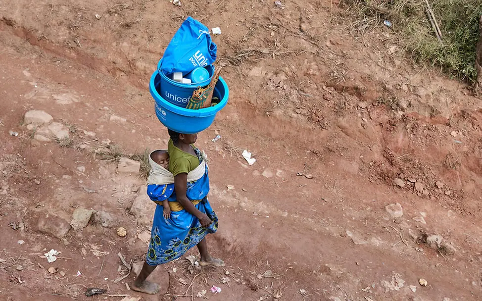 Mit Waschschüsseln, Seife und Hygieneartikeln hilft UNICEF Menschen in Krisenregionen, die Verbreitung von Krankheiten zu vermeiden. | © UNICEF/Andriantsoarana Was ist ein Tsunami: Eine Frau mit einem Baby auf dem Rücken trägt ein Hygiene-Set von UNICEF auf dem Kopf.