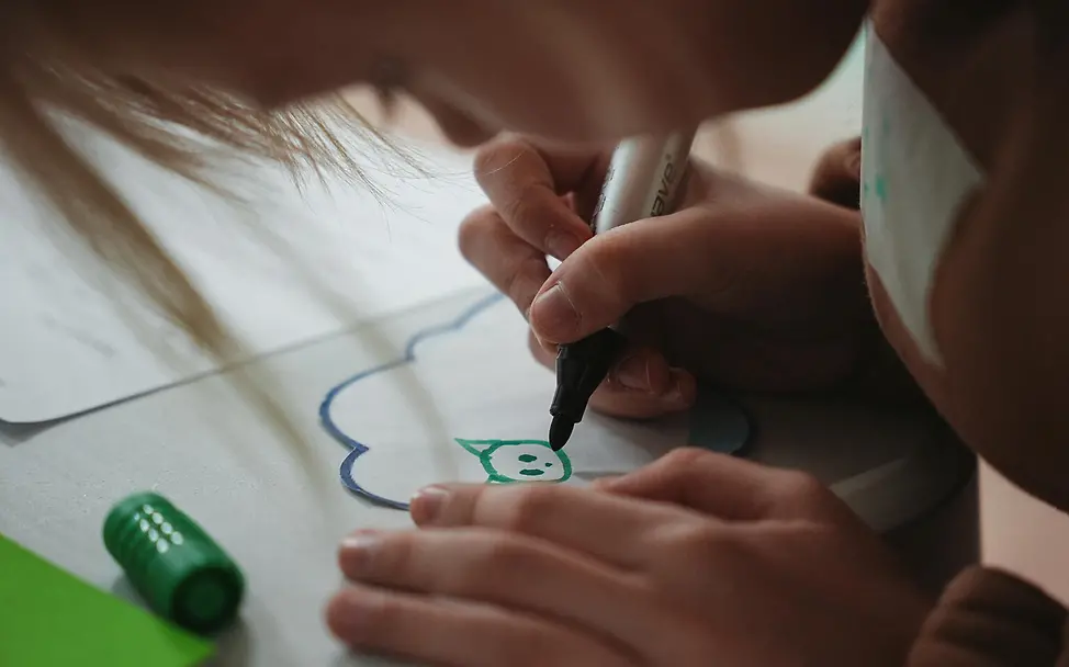 Ukraine: Kind bei einer Zeichenaufgabe | © UNICEF/Kruchkova Ukraine: Ein Kind zeichnet konzentriert auf Papier; sichtbar sind die Hände, ein Filzstift und ein kleiner grüner Deckel.