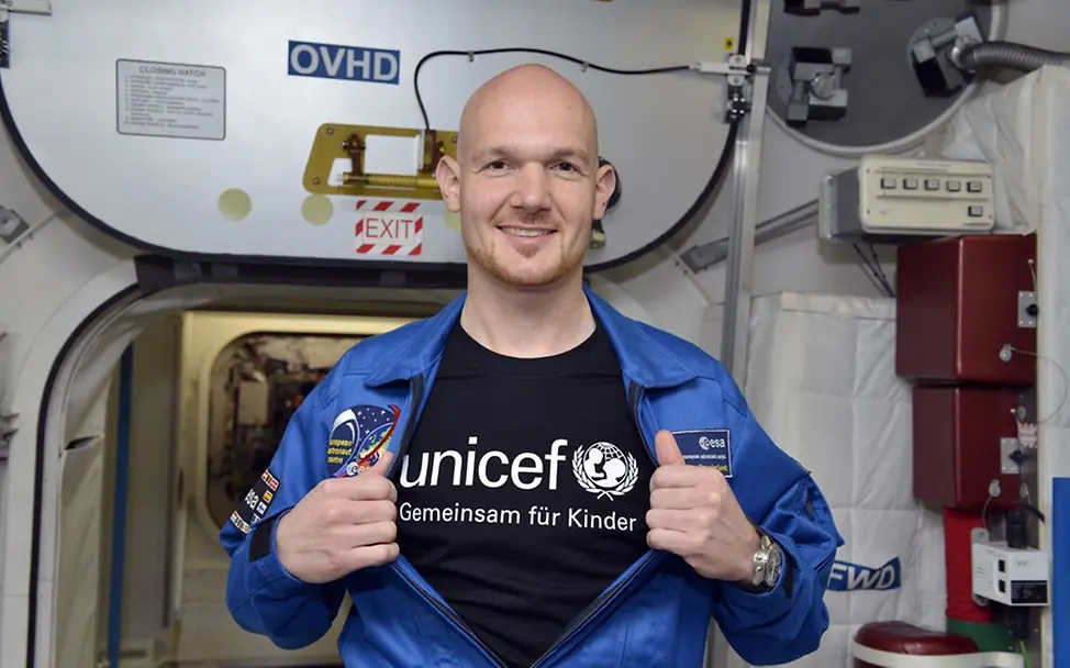 Alexander Gerst im All mit UNICEF | © UNICEF DT/Hyou Vielz Der Astronaut Alexander Gerst steht in einem Raumschiff und öffnet seine Jacke, worunter ein UNICEF T-Shirt zum Vorschein kommt.