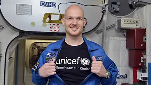 Der Astronaut Alexander Gerst steht in einem Raumschiff und öffnet seine Jacke, worunter ein UNICEF T-Shirt zum Vorschein kommt.