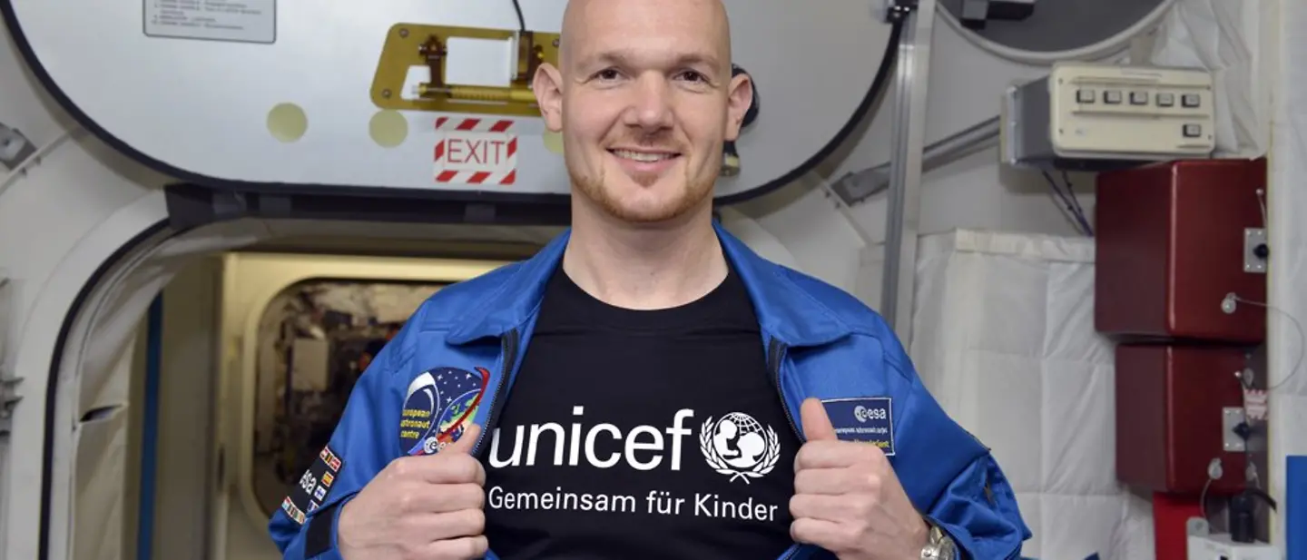 Alexander Gerst im All mit UNICEF | © UNICEF DT/Hyou Vielz Der Astronaut Alexander Gerst steht in einem Raumschiff und öffnet seine Jacke, worunter ein UNICEF T-Shirt zum Vorschein kommt.