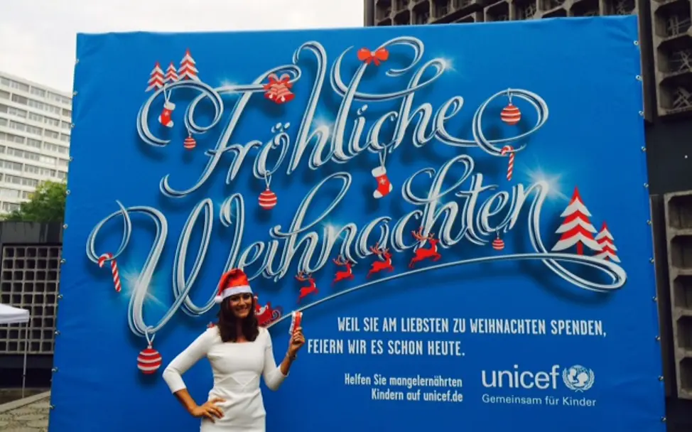 Sandra Thier präsentiert Weihnachten im Sommer | © UNICEF DT UNICEF-Weihnachten im Sommer: Sandra Thier vor Plakat