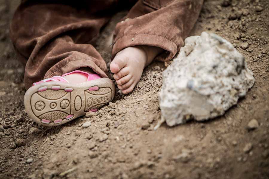 Siad sitzt im Dreck und spielt, während seine Familie auf Essensgutschriften in einem Flüchtlingscamp im Irak wartet. | © UNICEF/Schermbrucker Ein Schuh fehlt dem Kind, das auf der Erde sitzt.