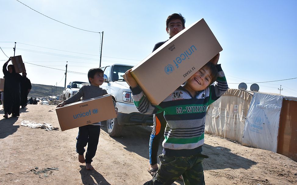 Drei Geschwister tragen in einem Flüchtlingslager im Irak Hilfspakete von UNICEF, die sie gerade erhalten haben.
