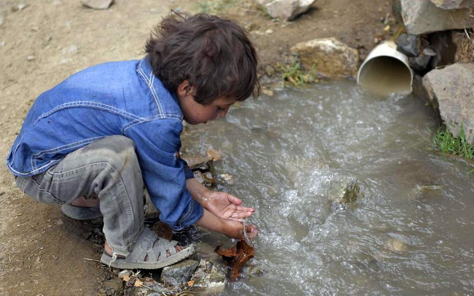 Kind schöpft mit den Händen Wasser neben einem Abwasserrohr und riskiert damit eine Choleraerkrankung.