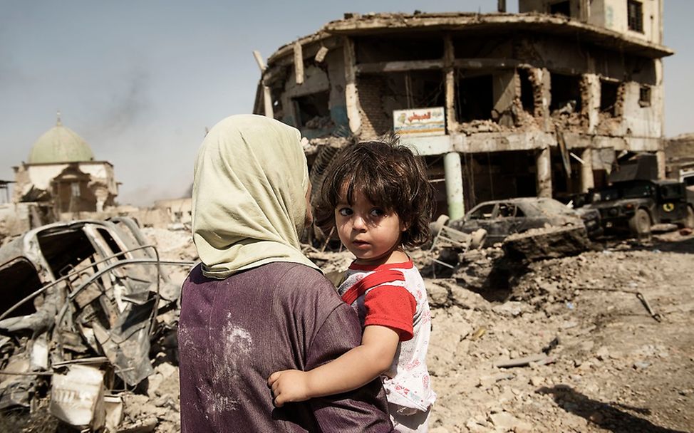 Kind auf dem Arm einer Frau vor den Trümmern eines Hauses im Iraq