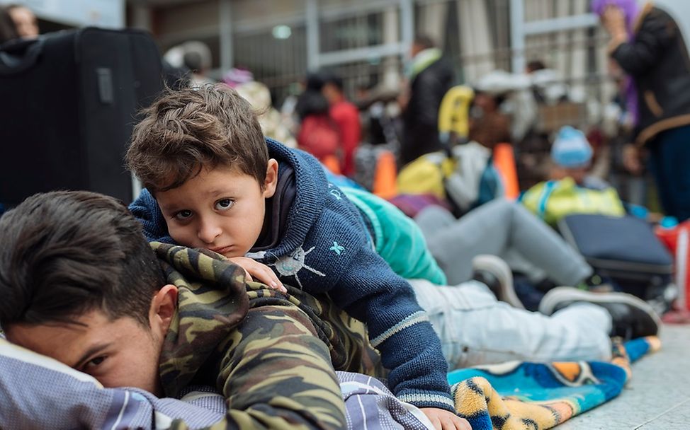Ein Kind liegt auf dem Rücken seines großen Bruders in einem Saal wartender Migranten