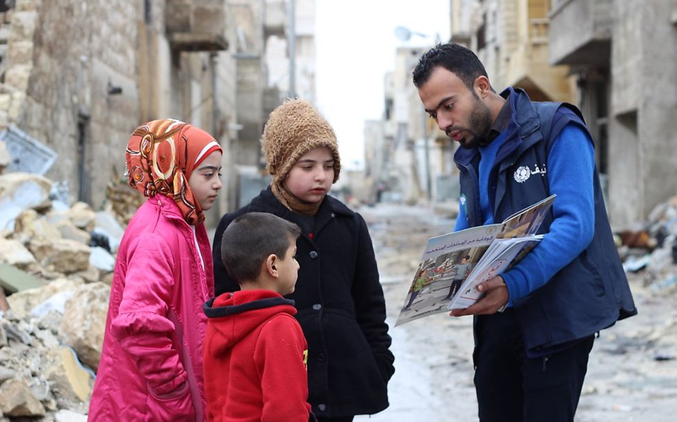 Ein UNICEF Mitarbeiter klärt drei Kinder über die Risiken in der vom Krieg zerstörten Umgebung auf.