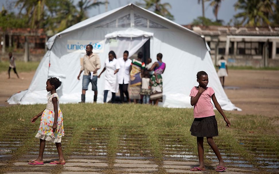 Zwei Kinder im Vordergrund laufen an einem UNICEF-Zelt im Hintergrund vorbei, aus dem zwei Ärzte kommen.