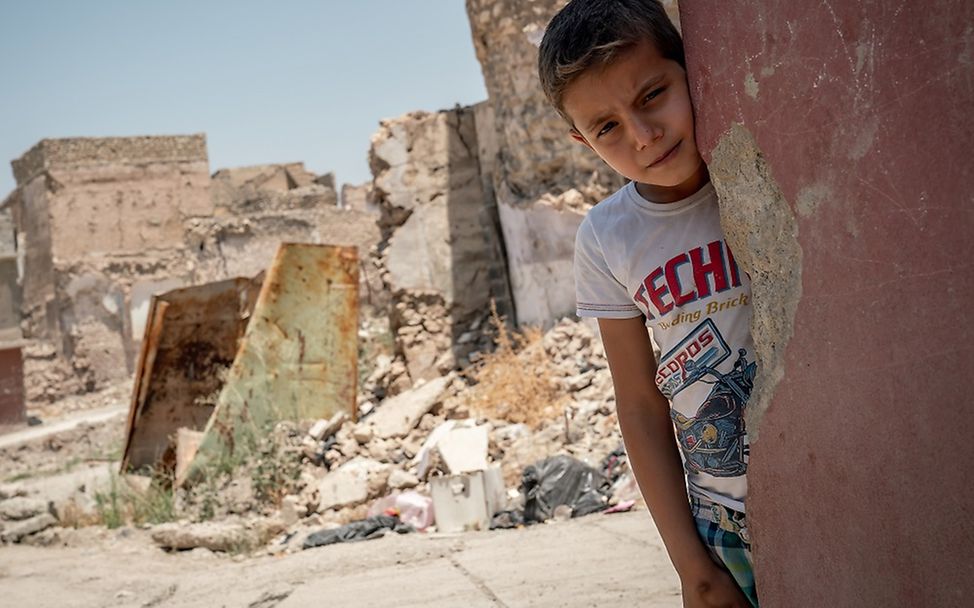 Irak: Der Junge Ahmad steht angelehnt an einer Hausecke mit Einschussloch. Im Hintergrund Schutt und Geröll.