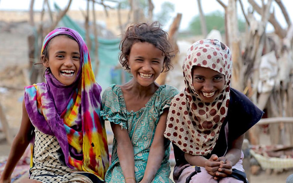 Wohlergehen von Kindern weltweit fördern: Drei Mädchen sitzen lachend auf einer Mauer.