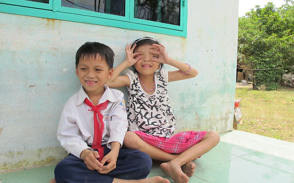 Vietnam: Ly sitzt neben ihrem Bruder und formt mit ihren Händen eine Kamera.