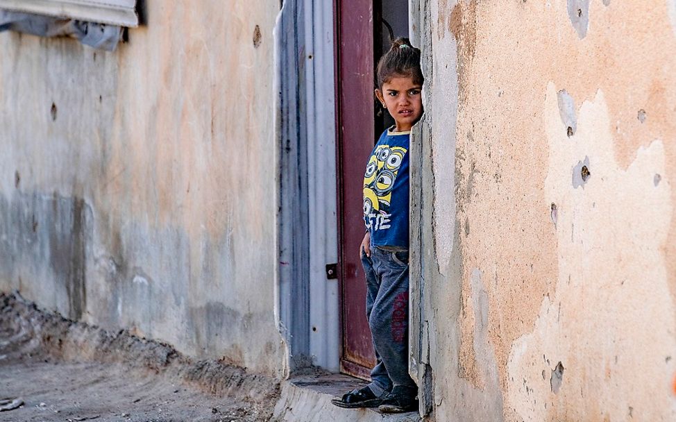 Syrien: Ein Mädchen steht in einer Haustür. Die Wand des Hauses ist gezeichnet von Einschusslöchern. 