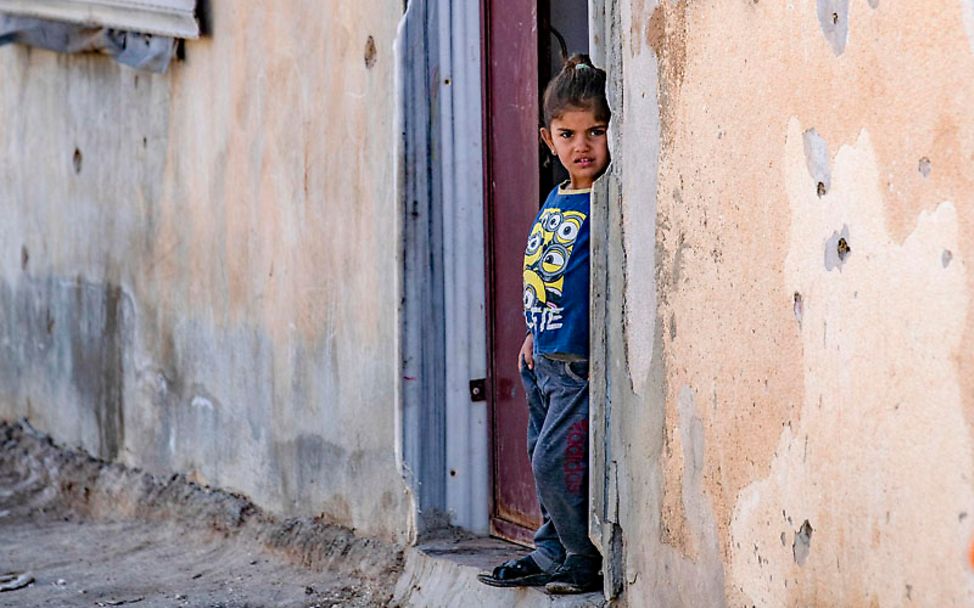 Syrien: Ein Mädchen steht in einer Haustür. Die Wand des Hauses ist gezeichnet von Einschusslöchern