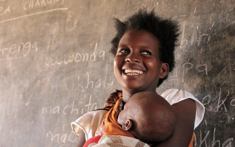 Schulen für Afrika: Eine junge afrikanische Frau steht mit ihrem Baby auf dem Arm in einem Klassenzimmer.