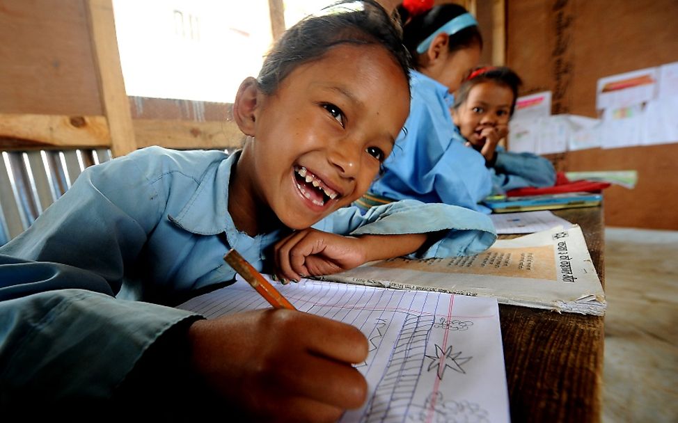 Nepal: Ein lachendes Mädchen sitzt an einem Pult und schreibt in ein Heft.