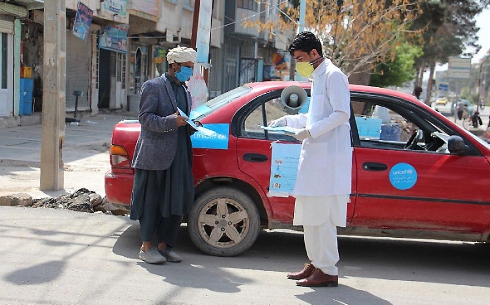 Afghanistan: Vor einem Auto stehen zwei Männer mit Mundschutz.
