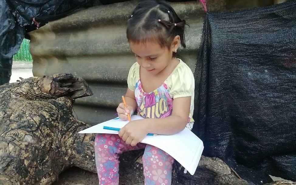Covid-19: In Kolumbien sitzt ein Mädchen sitzt auf einem Baumstamm und schreibt in ein Schulheft.