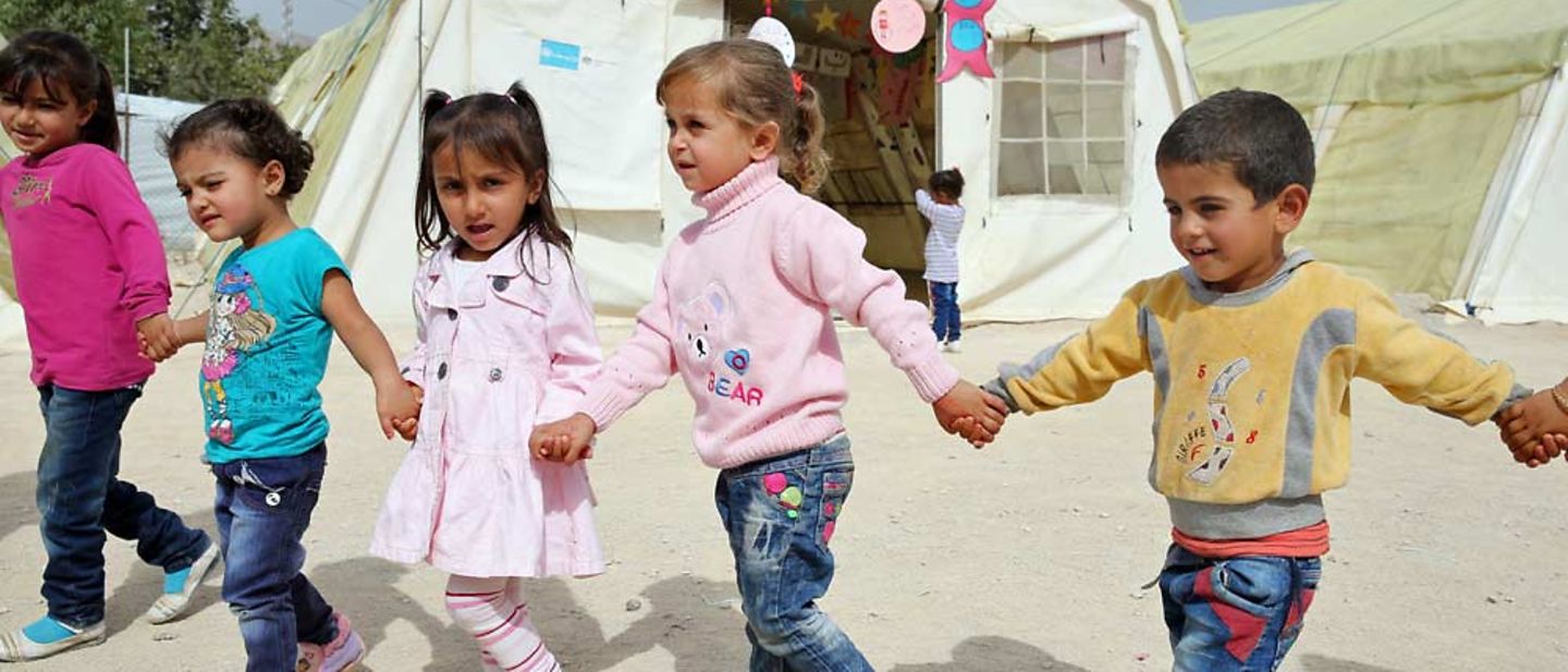 Mehrere Kinder spielen Hand in Hand in einer Flüchtlingsunterkunft vor einem UNICEF-Zelt.
