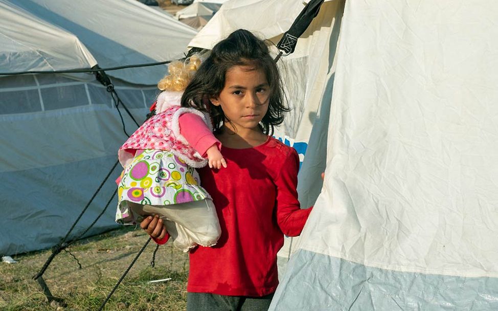 Ein Flüchtlingsmädchen steht mit einer Puppe auf ihrem Arm vor einem der Zelte im neuen Lager.
